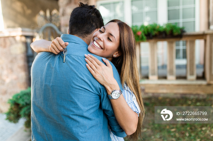 Happy young couple with key standing and hugging outside in front of their new home.