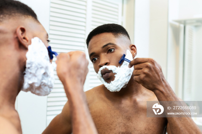 African american man shaving in bathroom, looking at mirror