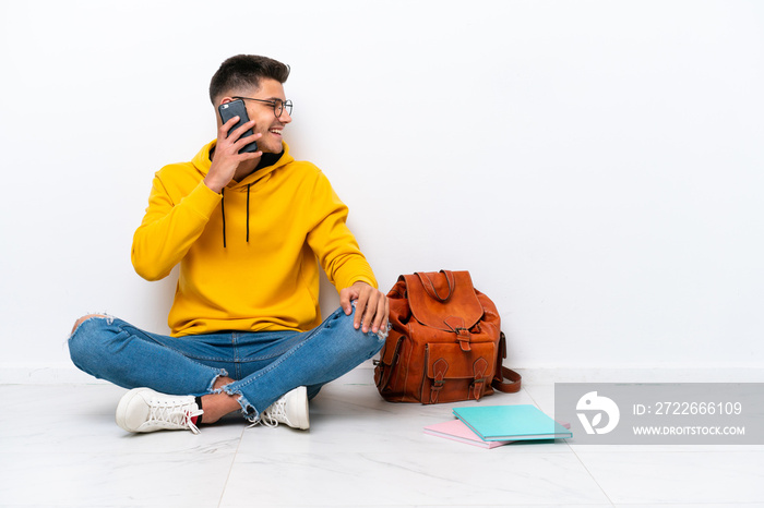 Young student caucasian man sitting one the floor isolated on white background keeping a conversation with the mobile phone with someone