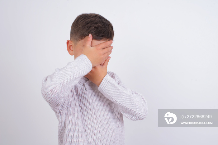 Caucasian kid boy wearing knitted sweater against white wall Covering eyes and mouth with hands, surprised and shocked. Hiding emotions.