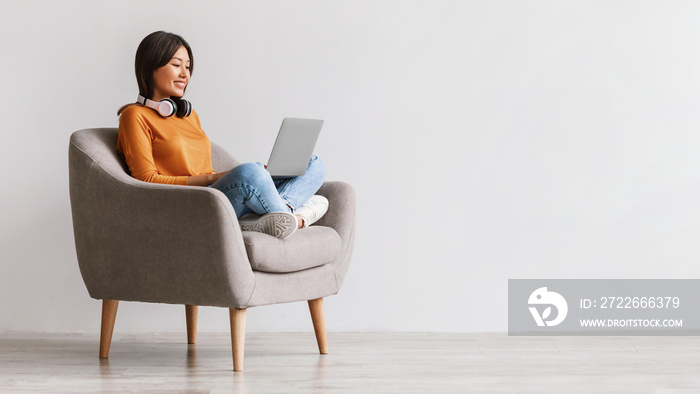 Asian woman sitting in armchair with laptop and headphones, studying online, having remote job, video conferencing