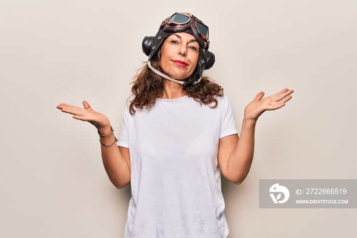 Middle age brunette woman wearing goggles and retro aviator leather hat over white background clueless and confused with open arms, no idea and doubtful face.