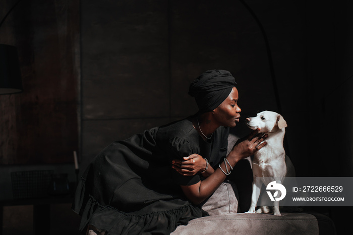 Young African woman in black turban and dress sits on cozy chair with little white dog strokes puppy at home. Owners and pets. Domestic animals. Pretty Brazilian girl taking care of hound. Mockup