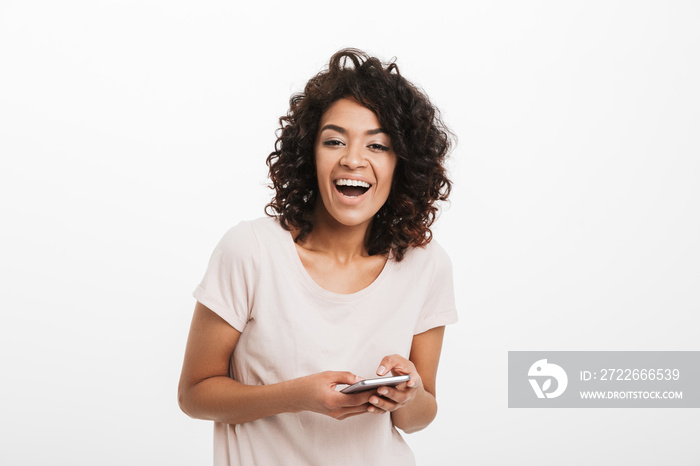 American woman 20s with curly hair holding mobile phone and smiling on camera, isolated over white background