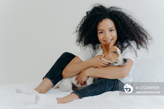 Female pet owner poses in clean white bedroom, hugs dog, plays with best friend, wears casual clothing, has cheerful expression