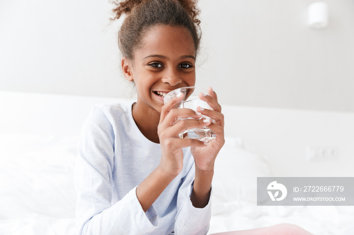 Image of happy african american little girl drinking water in bed at home