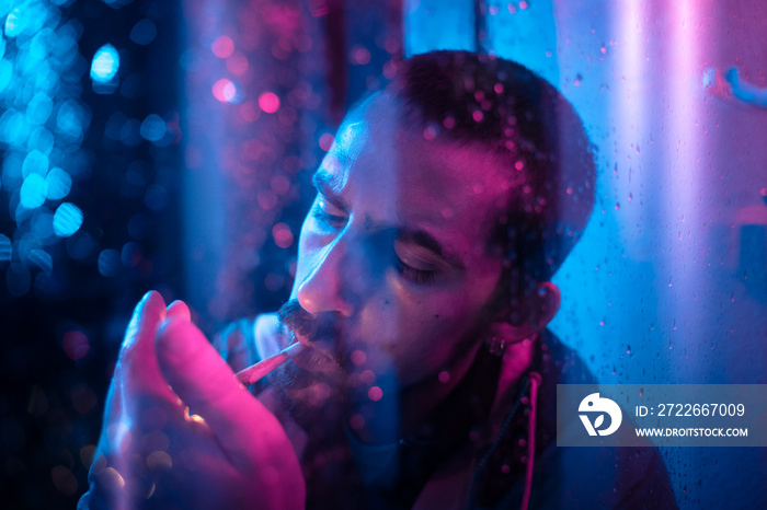 Beautiful cinematic portrait of stylish man in neon lighted window with rain drops. Bright neoned colors. Caucasian model, musician outdoors. Youth culture, scary mist style and music concept.