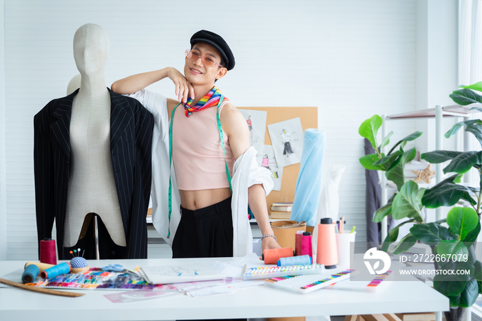 Portrait of happy young Asian LGBTQ gay dressmaker standing confident pose with mannequin behind working desk, sketch of fashion dress design, tailor tools in studio, small business owner concept