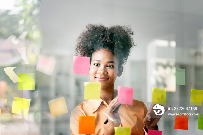 Young smiley attractive, businesswoman using sticky notes in glass wall to writing strategy business plan to development grow to success