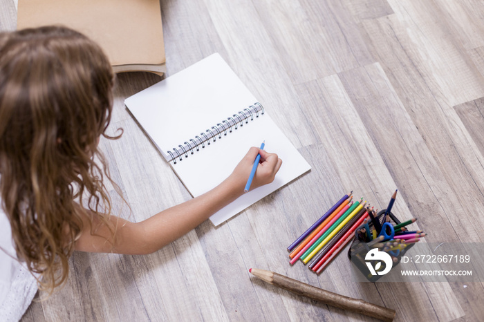 Cute schoolgirl sitting on the floor and drawing on a notebook. brown wood background. Back to school concept. Pencils and notebook besides. top view