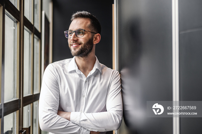 Young smiling attractive elegant bearded businessman standing near window with arms crossed and looking trough it.