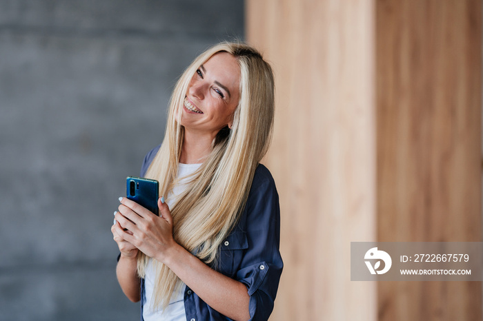 Beautiful shy blonde laughing swedish girl in grey shirt holding phone toothy smiling indoors. Attractive Caucasian young woman enjoying new cellphone. Electronics and gadgets.