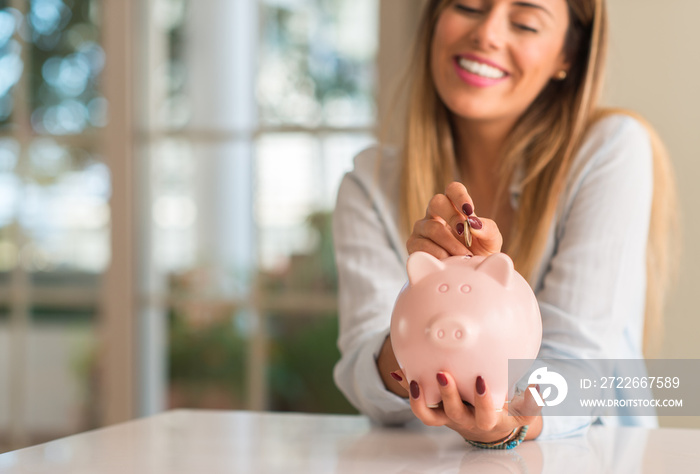 Beautiful young woman smiling holding a coin investing in to a piggy bank at home. Business concept.