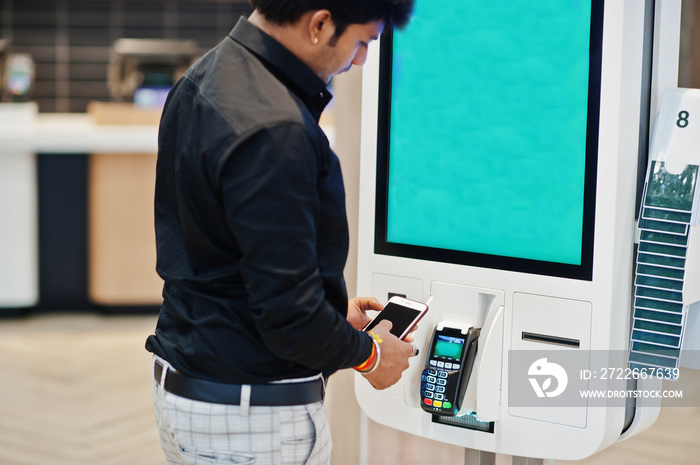 Indian man customer at store place orders and pay by contactless credit card on mobile phone through self pay floor kiosk for fast food, payment terminal. Pay pass.