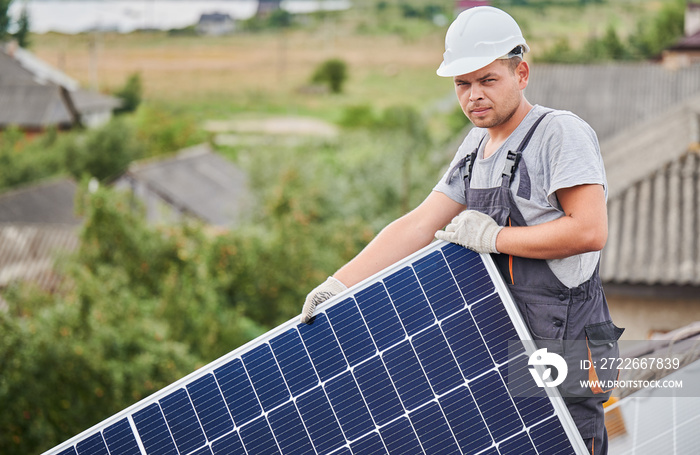 Portrait of man worker mounting photovoltaic solar moduls on roof of house. Electrician in helmet installing solar panel system outdoors. Concept of alternative and renewable energy.