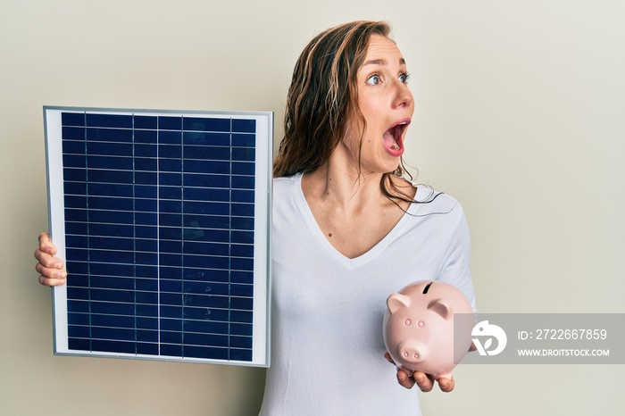 Young blonde woman holding photovoltaic solar panel and piggy bank angry and mad screaming frustrated and furious, shouting with anger. rage and aggressive concept.