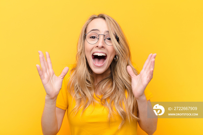 young pretty blonde woman feeling shocked and excited, laughing, amazed and happy because of an unexpected surprise against flat color wall