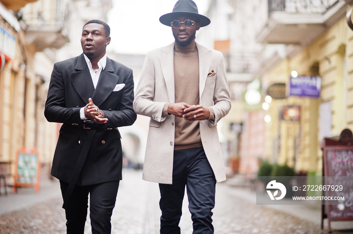 Two fashion black men walking on street. Fashionable portrait of african american male models. Wear suit, coat and hat.