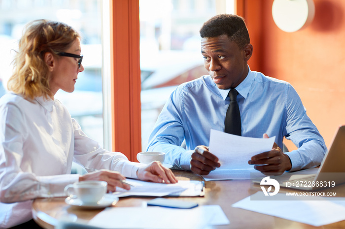 Adult professional diverse man and woman having business meeting and talking while sitting with papers at table