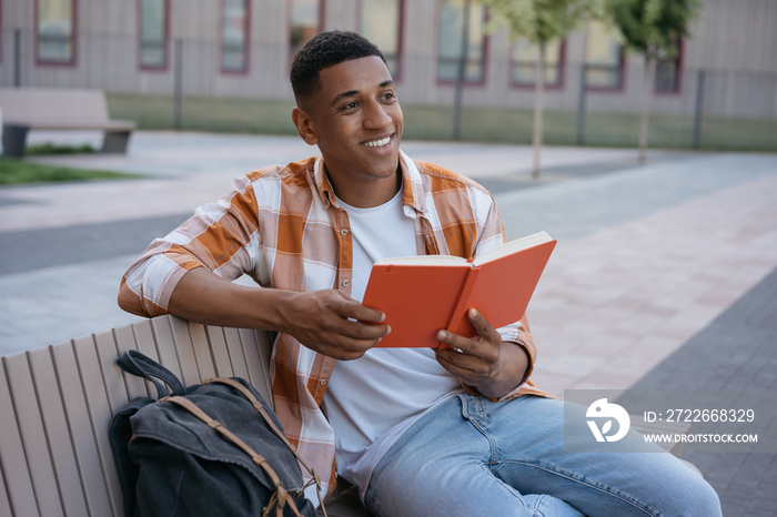 Smiling African American student studying, learning language sitting in university campus, education concept. Young handsome man reading book, relaxing in park