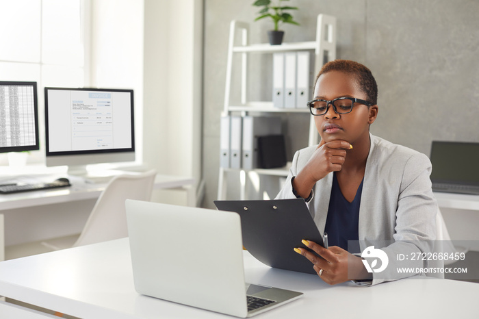 Black woman reading documents and thinking sitting at office desk with modern laptop. Business lady in glasses considering question, studying contract terms and conditions, making important decision