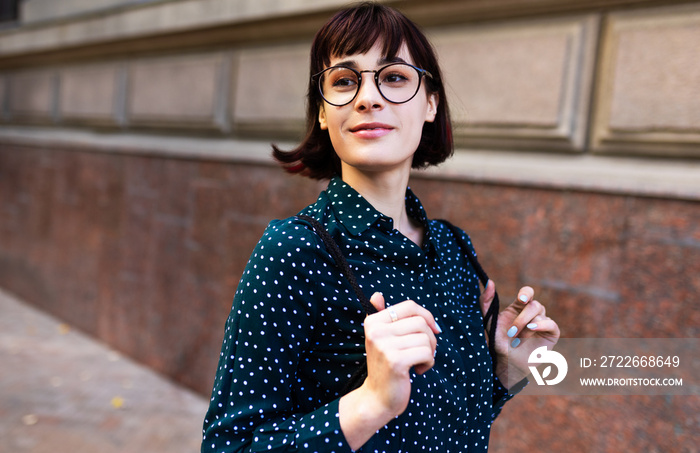 Young woman wearing transparent eyeglasses with backpack going to the office. Young student female walking beside building campus in the street.