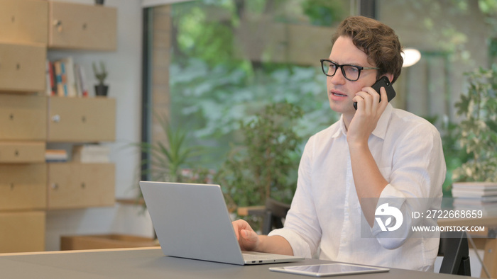 Young Man Talking on Smartphone while Using Laptop in Modern Office