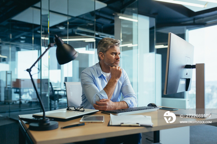 A gray-haired man thinks about the tasks, a businessman with a beard works in a modern office at the computer,