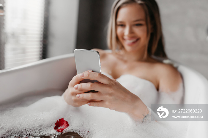 Cheerful woman using smartphone while lying in foamy bath, reading beauty blog or watching video online and smiling