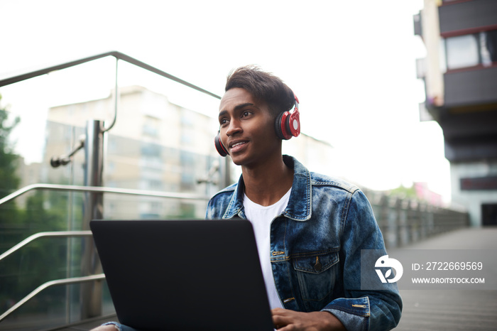 Stylish hindu teenager listening audio lesson of learning languages in modern headphones sitting in urban setting with computer connected to 4G internet.Blogger enjoying music from radio website