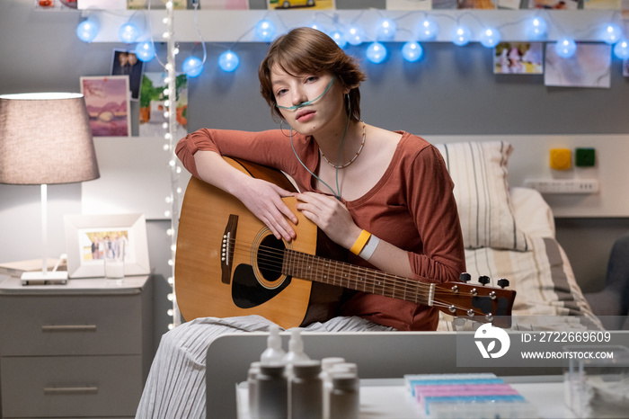 Teenage female patient with acoustic guitar sitting on bed in modern hospital ward and looking at camera