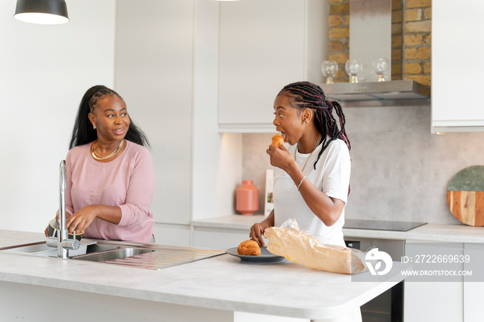 Young woman washing dishes while girlfriend eating croissant in kitchen