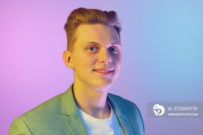 Young handsome man with stylish hairdo in jacket posing, looking at camera and smiling over purple background in neon light. Concept of emotions, lifestyle, youth, modern fashion. Ad