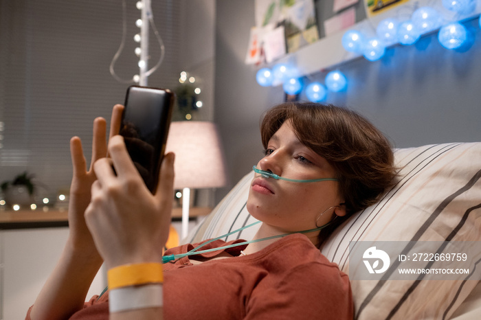 Brunette teenager with oxygen nose pipe texting in smartphone while lying on bed in hospital ward at leisure