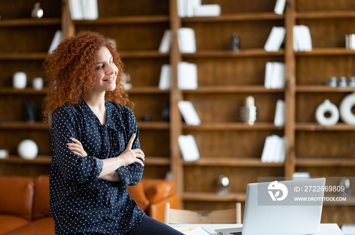 Contemporary woman entrepreneur stands with arms crossed in the office, cheerful freelancer woman in informal wear leaned at the desk and looks away, inspired small business owner