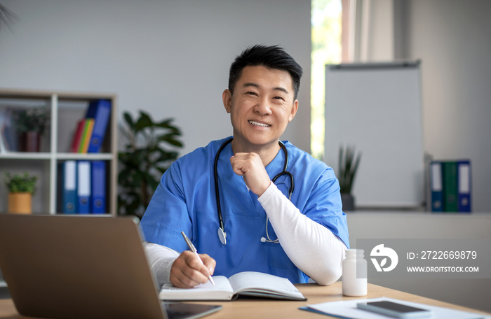 Cheerful young korean male doctor in uniform with stethoscope at workplace work with laptop