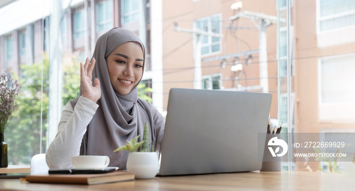 smiling beautiful muslim businesswoman using a laptop for online video meeting sitting at the desk in modern office space, looks at the webcam and waving hello