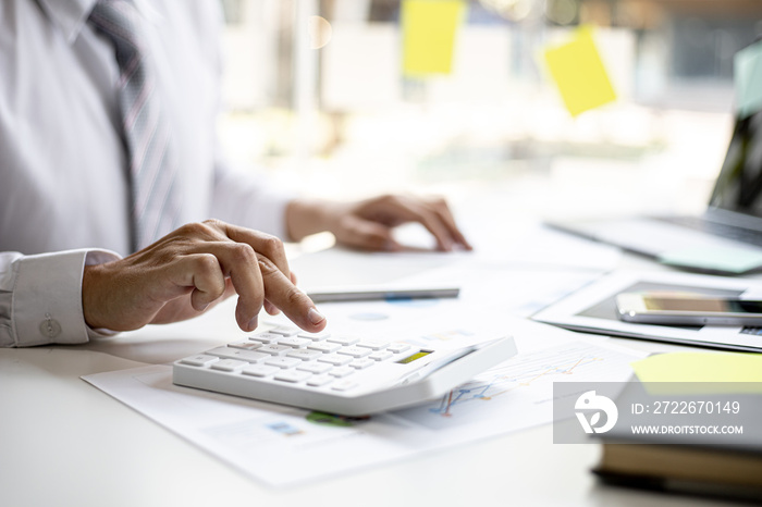 A businessman is using a white calculator to calculate financial numbers, he is sitting in his private office, the businessman examines the financial data from the corporate finance chart.