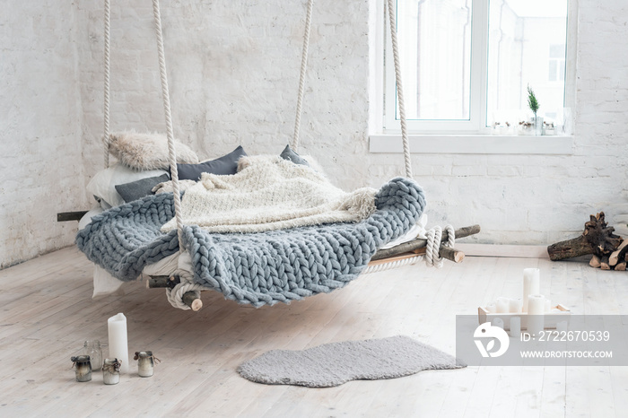 White loft interior in classic scandinavian style. Hanging bed suspended from the ceiling. Cozy large folded gray plaid, giant knit blanket, super chunky yarn, arm knitting. Trendy room design.