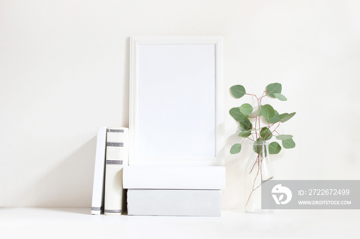 White blank wooden frame mockup with a green eucalyptus branches in glass bottle and pile of books lying on the table. Poster product design. Styled stock feminine photography. Home decor.