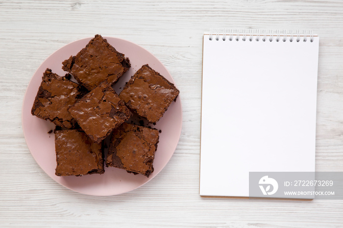 Homemade chocolate brownies on a pink plate, blank notepad, top view. Flat lay, overhead, from above. Copy space.