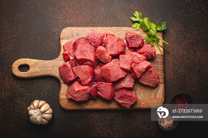 Raw beef meat chopped in cubes with bunch of fresh parsley, garlic, salt and pepper on wooden cutting board for cooking stew or meat dish on brown dark stone concrete background top view flat lay