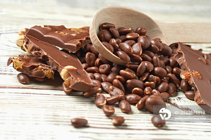 Lot of dragees with glazed peanuts strewn from a wooden spoon near broken chocolate bar with caramel, raisin, big nuts on old worn white rustic table. Close-up