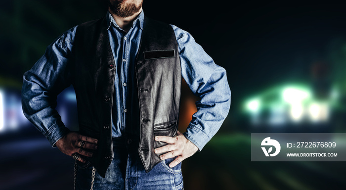 Photo of man in jeans shirt and leather biker vest standing on neon night city background.