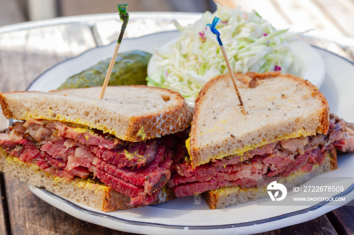 Pastrami sandwich sliced in half on rye bread with mustard on a white plate on a wood table as seen from above. Coleslaw and dill pickle on side. Toothpicks in both halves of sandwich.