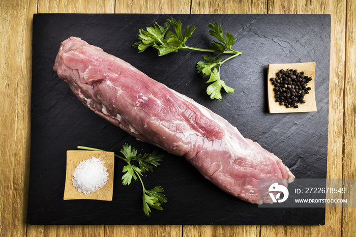 pork tenderloin next to salt, black pepper and parsley on a black slate plate on a wooden table