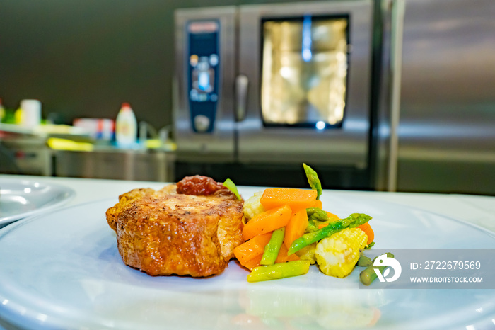 Pork steak with side dish. Serving dishes in a restaurant. Piece of meat on a plate with a vegetable side dish. Book of recipes. Dish with pork on the background of a combi oven. Garnish of carrots.