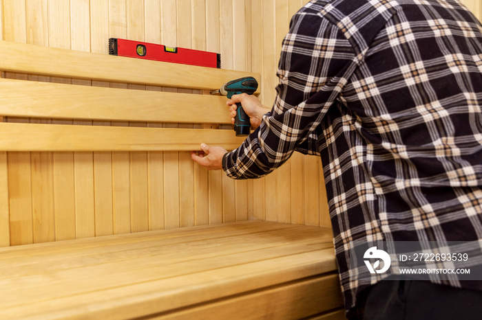 sauna construction - man screwing wooden bench backrest on the wall