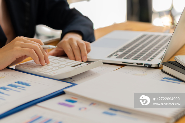 An Asian accountant’s hand is pressing a calculator and simultaneously analyzing financial graphs and a laptop next to it while still holding a pen in his finger.