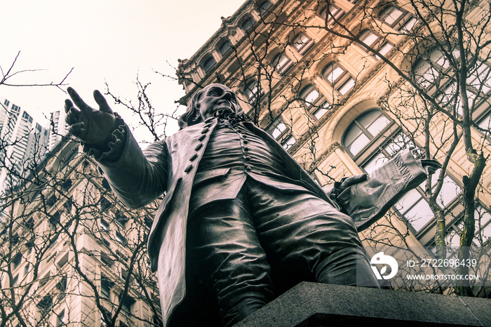 Upward view of a statue of Benjamin Franklin with tree branches and buildings on background.